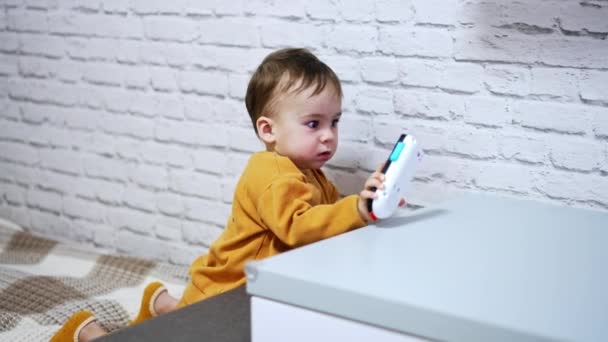 素晴らしいコーカサス人の赤ん坊の男の子は屋内ベッドの上に座っています おもちゃの電話で遊んでいる穏やかな愛らしい幼児 — ストック動画