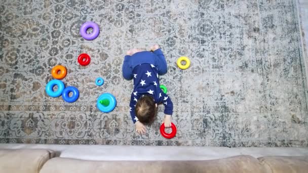 小さな男の子が部屋の床にひざまずいている おもちゃのリングが散りばめられています 子供はカメラを見ていると頭を上げる トップビュー — ストック動画