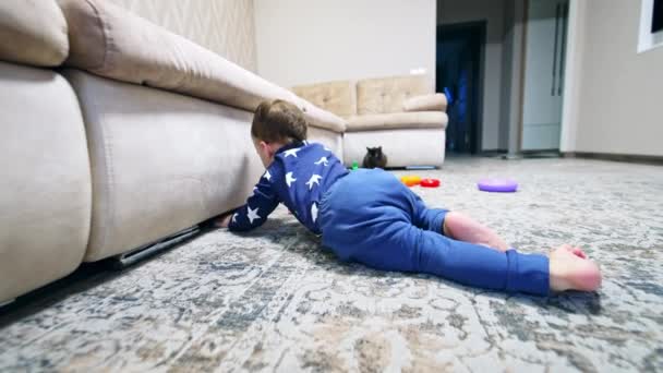 青い服を着ているトッドラーはソファーの下を見下ろす床に横たわっている ベイビーはソファーの下から電話を引っ張ってからプッシュします — ストック動画
