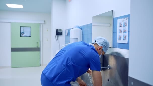 青い制服の男性医師 マスクと帽子は慎重に手を洗っています 手術前の部屋で手術の準備をする外科医 手術における不純物 — ストック動画