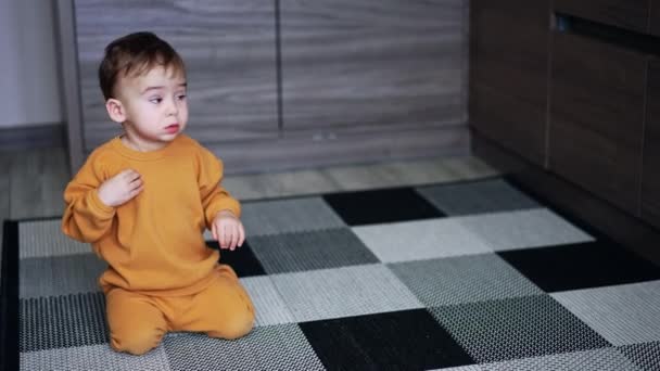 オレンジ色の服の美しい幼児はキッチンフロアに座っています 赤ちゃんはドアを開けようとしているクローゼットにクロールアップ — ストック動画