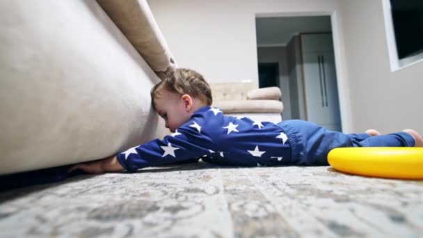 小孩把电话从沙发上拿开 身穿蓝色衣服的男婴躺在地板上 手伸向家具下面 — 图库视频影像