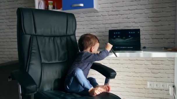 小孩坐在一个大皮椅上 婴儿转动椅子把遥控器放在桌子上 — 图库视频影像