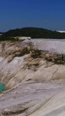Bir kaolin madeninin tepeciği ve eğimli bölgesinde çiğ kaolin üretiliyor. Gökyüzünü yansıtan küçük mavi bir göl. Yeşil orman arka planı. Dikey video