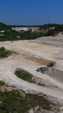 Kaolin kil madeninin gelişmemiş toprakları. Kazı makinesini yükleyen kazıcıyla beyaz manzara. Yeşil ağaçlar ve açık gökyüzü arkaplanı. Dikey video