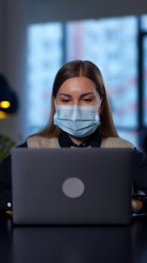 Bilgisayarına bir şey yazan maske takan kadın bir ofis çalışanı. Bayan daha iyi görebilmek için ekrana yaklaşıyor. Ofis geçmişi bulanık. Dikey video