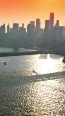 Güzel köprülerin altında East River 'dan geçen tekneler. Batan güneş ışınlarında ünlü New York gökdelenlerinin siluetleri. Pembe turuncu gökyüzü arka planı. Dikey video