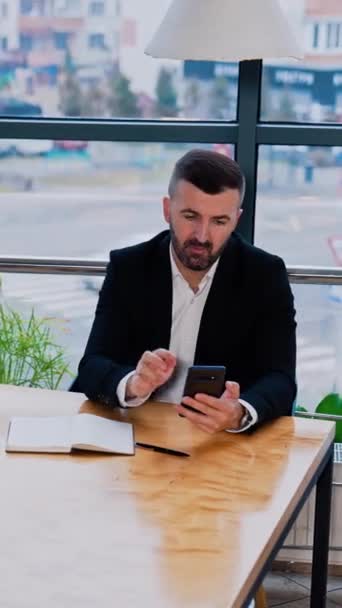 身穿黑色西服 留着胡子的男子坐在桌旁 商人使用电话搜索信息并显示搜索结果 垂直录像 — 图库视频影像