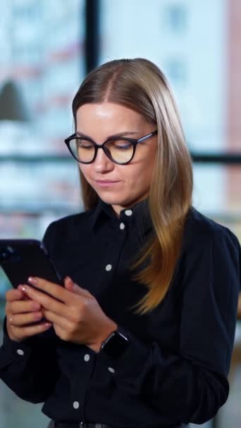 办公室里的金发女人想打个电话 戴眼镜的女士拿着智能手机在她的耳朵等待答案 模糊的背景 垂直录像 — 图库视频影像