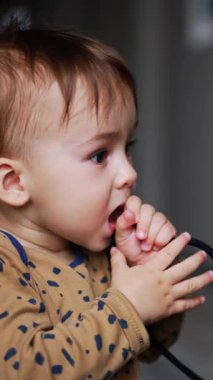 Küçük bebek ayakkabı bağcığıyla oynamakla meşgul. Komik çocuk ağzını kocaman açıyor ve bir kordon alıp ağzına giriyor. Kapatın. Arkaplan bulanık. Dikey video