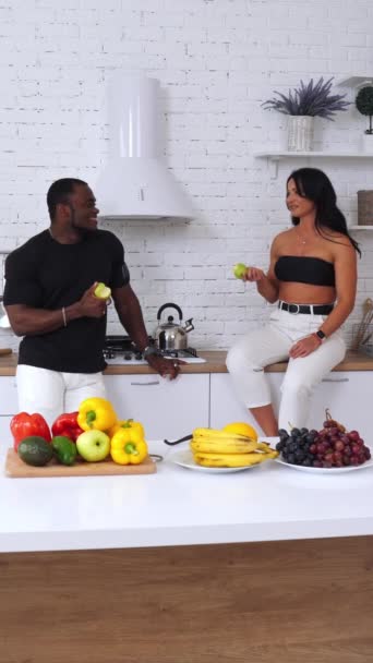 キッチンでリンゴを食べたり 会話をするカップルにぴったりです 健康的な有機食品でいっぱいのテーブルの近くで話す陽気な人々 バーティカルビデオ — ストック動画