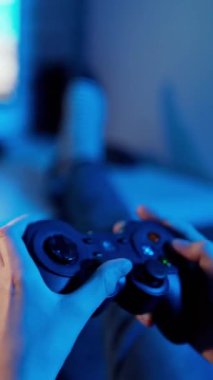 Oyun oynayan genç bir oyuncu kumanda kullanıyor. Playstation ekranının önündeki oğlanın elinde joystick var. Yakın plan. Mavi ışık. Dikey video
