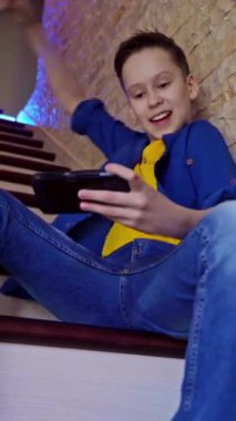 Evdeki merdivenlerde oturan bir çocuğun manzarası. Cep telefonuyla oynayan mutlu genç oyunu kazanıyor. Oyun konsepti. Dikey video