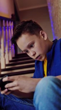 Telefondaki oyunlara bağımlı bir genç. Çocuk evde merdivenlerde oturuyor ve cep telefonuyla video oyunları oynuyor. Dikey video