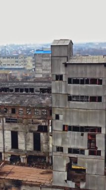 Şehir binalarının arka planında gri duvarları ve kırık camları olan büyük bir devlet fabrikası. Sanayi bölgesi. Hava görüntüsü. Dikey video