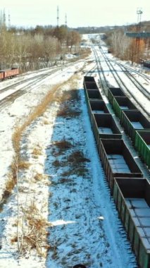 Bir yük treni, kışın kentin eteklerinde sayısız sıra konteynır bulunan tren istasyonunun arka planındaki demiryolu üzerinde konteynırlarla dolu bir treyleri çeker. Hava görüntüsü. Dikey video