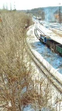 Konteynırları olan bir yük treni bir kış günü raylar boyunca şehre doğru hareket eder. Malların nakliyesi. Hava görüntüsü. Dikey video
