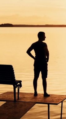 Bir adam köprüde duruyordu ve sonra bir bankta oturmuş nehrin arka planındaki doğanın güzelliğine ve bir yaz akşamında gün batımına bakıyordu. Dikey video
