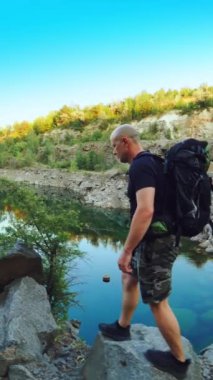 Yazlık giysiler içindeki bir turist elinde taşlarla bir taş ocağının kenarında yürüyor ve etrafındaki güzel göle ve doğaya bakıyor. Güzel manzara. Dikey video