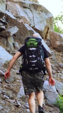 Sırt çantalı bir gezgin sıcak havada öğleden sonra taşlarla dağın tepesine yükselir. Turizm. Dikey video