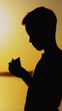 Genç çocuk, gün batımında nehrin kenarında duran kağıt bir gemi yapıyor. Dikey video dışında kağıt tekneyle oynayan çocuğun silueti.