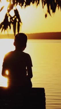 Küçük çocuk akşamleyin nehirde ahşap bir rıhtımda oturuyor. Gün batımında nehre bakan bir çocuğun fotoğrafı. Yazın dikey videoda birini ya da bir şeyi bulmaya çalışıyor.
