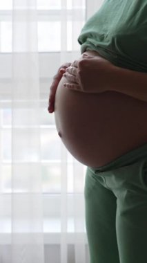 Koca göbekli bir kadın büyük ışıklı pencereye yaklaşır. Müstakbel anne nazikçe ve sevgiyle karnını okşuyor. Dikey video
