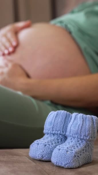 一个大肚皮的孕妇坐在沙发上 女士温柔地爱抚她怀孕的肚子 然后把婴儿袜子放在上面 垂直录像 — 图库视频影像