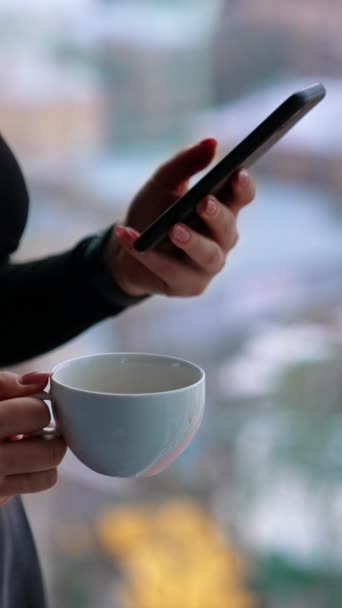 美丽的女性 站在窗边 一边喝着一杯咖啡一边沉思地浏览智能手机 — 图库视频影像