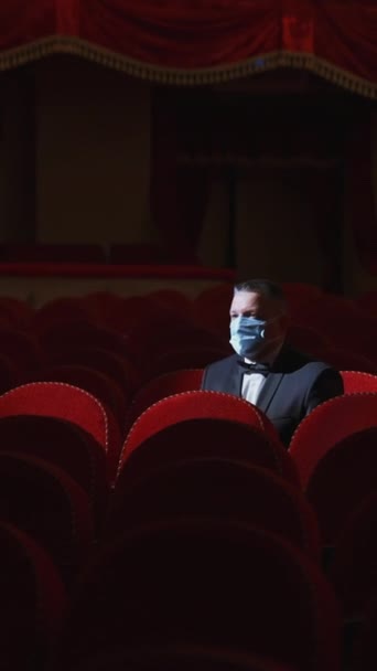 金持ちは空っぽの赤い椅子の列の中にいる 暗い劇場の背景に一人で快適な椅子に座っている医療マスクのビジネスマン 世界のパンデミック バーティカルビデオ — ストック動画