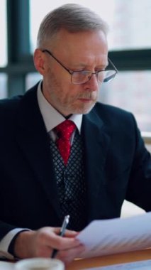 Resmi kostümlü kıdemli iş adamının portresi. Olgun bir adam dizüstü bilgisayarın önünde oturuyor ve pencerenin yanındaki ofiste kağıtlarla çalışıyor. Dikey video