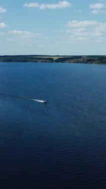 Doğa arasında geniş mavi bir nehrin panoramik görüntüsü. Yazın nehrin ortasında yüzen sürat teknesi. Hava görüntüsü. Dikey video