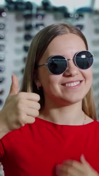 商店里戴时髦太阳镜的漂亮姑娘 在光学背景下 一个可爱的女孩用手在太阳镜上显示出好的标志的肖像 垂直录像 — 图库视频影像