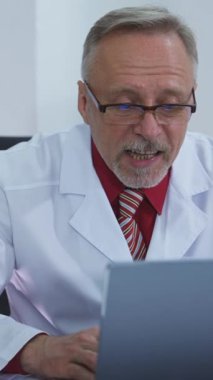 Uzaktan görüntülü arama yapan bir sağlık görevlisi. Beyaz önlüklü kıdemli doktor bilgisayarın önünde oturuyor ve tıp merkezindeki bir müşteriyle konuşuyor. Online danışmanlık. Dikey video