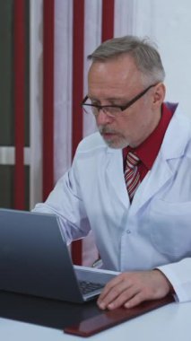 Yaşlı doktor laptopta video görüşmesi yapıyor. Üst düzey erkek doktor web sohbetinde web kamerasıyla iletişim kuran bilgisayar ekranına bakarken hastayla konuşuyor. Teletıp konsepti. Dikey video