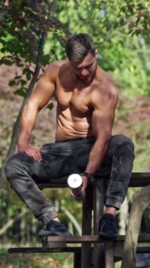 Sporcu doğada omuz kaslarını çalıştırıyor. Parkta oturan kaslı bir sporcu, dambılla kol kaslarını şişiriyor. Dikey video
