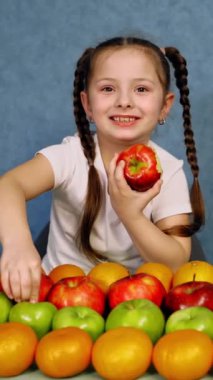 Küçük kız sulu elma yiyor. Masaya güzel yerleştirilmiş taze organik meyve. Mavi arka planda olgun meyveleri olan mutlu küçük kız. Dikey video