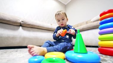 Oyuncakların yanında yerde oturan beyaz, tatlı bir oğlan. Piramitlerle oynayan sevimli bir çocuk. Düşük açı görünümü.