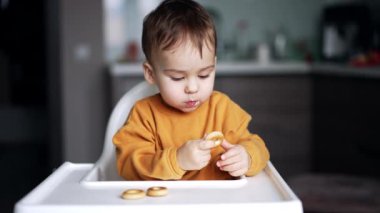 Yemek masasındaki sevimli çocuk ağzına simit alıyor. Evde bir bebeği beslemek. Bulanık arkaplan.