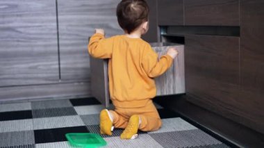 Turuncu takım elbiseli küçük bebek mutfaktaki sandığı açtı. Tanınmayan çocuk bulaşıkları çekmeceden çıkarıyor..
