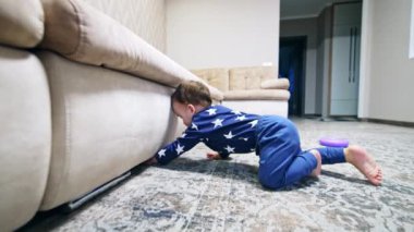 Bebek kanepenin altına girmeye çalışıyor. Çocuk yerde yatıp mobilyaların altına bakıyor. Çocuk ayağa kalkar ve oyuncaklarına gider..