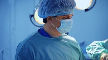 Şapkalı ve maskeli yetişkin bir erkek cerrah meslektaşıyla konuşuyor. Ameliyat halindeki bir doktorun portresi. Kapat..