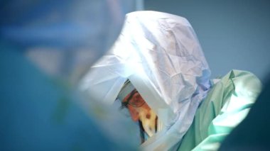 Gözlüklü ve koruyucu takım elbiseli bir cerrahın odaklanmış yüzü. Doktor operasyonda bir takımda çalışıyor. Bulanık arkaplan.