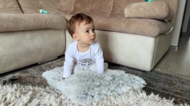 Beyaz elbiseli sevimli siyah saçlı erkek bebek yerde yumuşak yastıkla oynuyor. Çocuk kanepede ayakta duruyor..