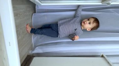 Beyaz tatlı beyaz çocuk balkon kapısında ve beyaz perdede duruyor. Meraklı çocuk kapıyı ileri geri hareket ettiriyor. Dikey ekran.