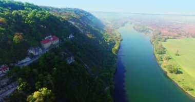 Dağların eteklerinde ağaçlarla kaplı huzurlu bir nehir. Dağda bulunan görkemli kaya manastırı. Hava görünümü.