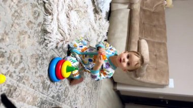 Rengârenk şortlu güzel beyaz erkek bebek yerde oturuyor. Çocuk oyuncakla oynuyor ve elini bir şey için kaldırıyor. Dikey video.