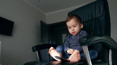 Küçük bebek televizyon kumandasıyla oynuyor. Siyah ofis koltuğunda oturuyor. Evde çıplak ayakla oynayan sakin bir çocuk. Düşük açı görünümü.