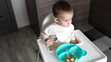 Beyaz gömlekli bebek mavi bir tabağın önünde oturuyor. Elinde kaşık tutan bir çocuk. Anne oğlunu besliyor. Üst görünüm.