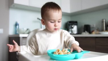 Küçük oğlan, önünde dolu bir tabakla yemek masasında oturuyor. Çocuk bir parçayı dikkatle alır ve ağzına çeker..